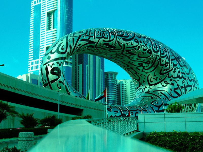 A modern architectural structure with Arabic calligraphy, surrounded by tall buildings and greenery.