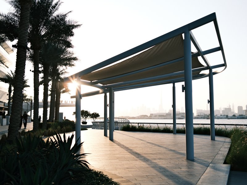 A modern pavilion with a sunshade, surrounded by palm trees and a waterfront view at sunset.