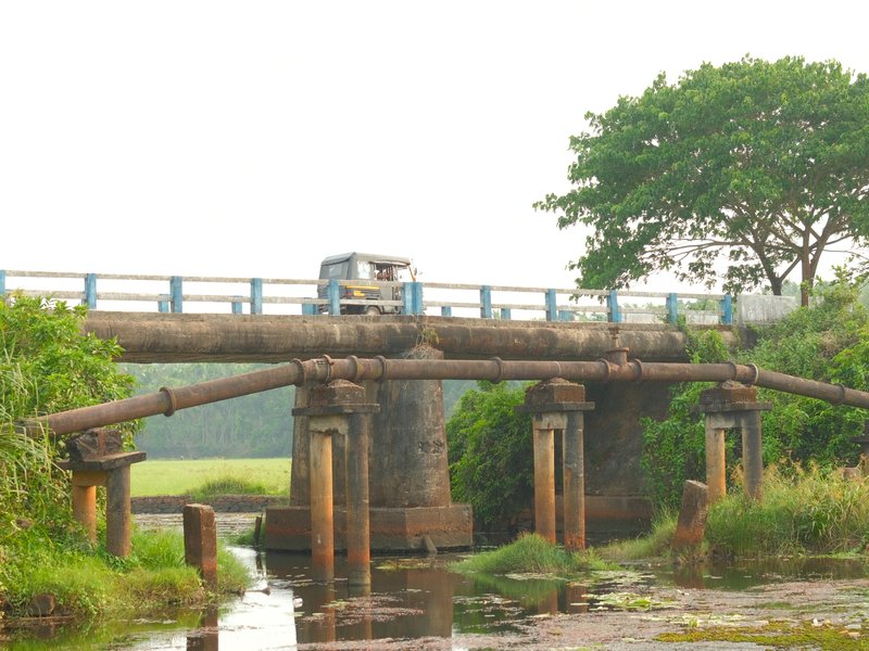 A weathered bridge over a small stream, surrounded by lush greenery and fields in the background.