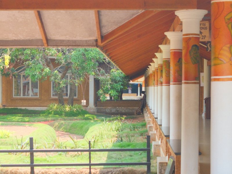 View of a corridor with decorative columns leading to a lush green garden and a building in the background.