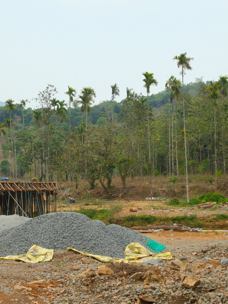 A construction site with piles of gravel and palm trees in the background under a clear sky.