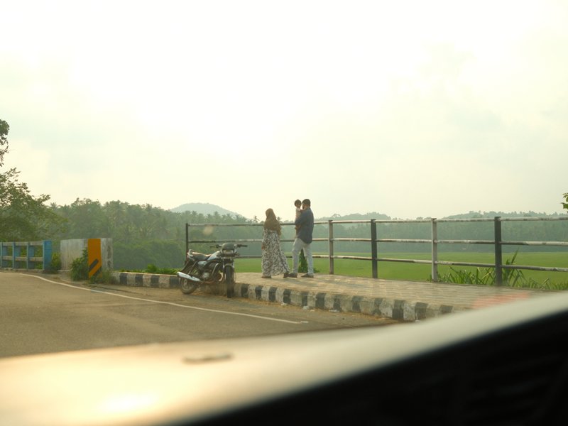 A couple walking along a path near a river, with greenery and hills in the background under a cloudy sky.