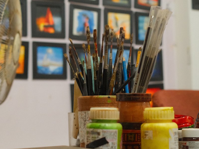 Close-up of paintbrushes and paint jars on a table with artwork in the background.