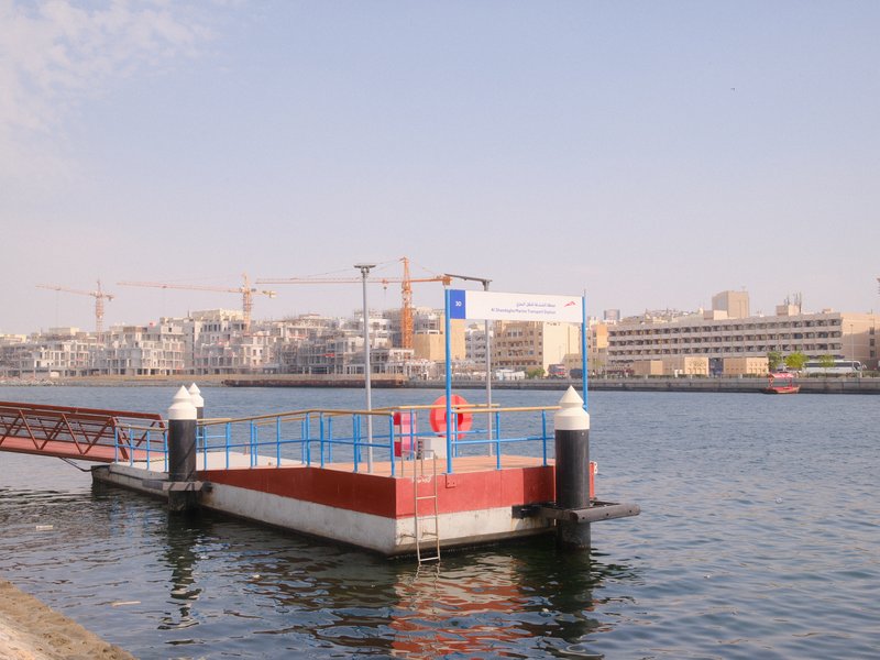 A waterfront scene featuring a dock with a ramp leading to a floating platform, surrounded by buildings.