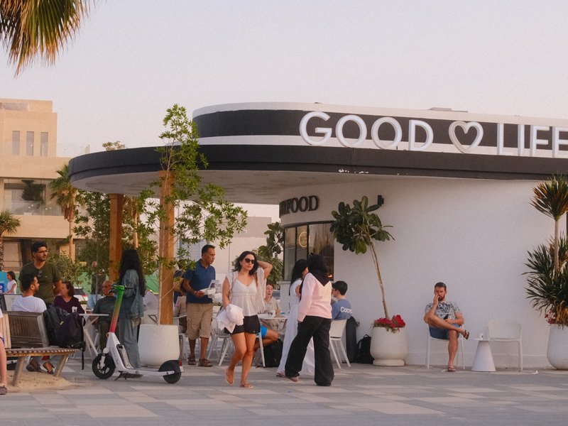 People socializing outside a modern café with a sign that reads 'GOOD LIFE' during sunset.