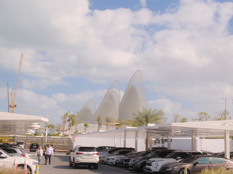 A modern architectural structure with curved shapes in the background, surrounded by parked cars and a clear sky.