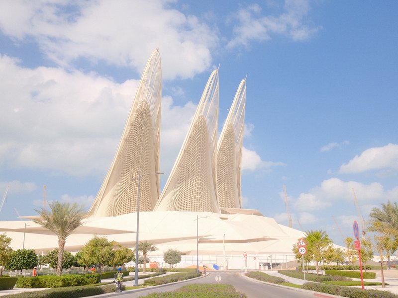Modern architectural structure with three sail-like towers against a blue sky and scattered clouds.