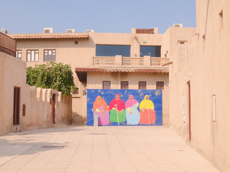 Colorful mural of four women in traditional attire on a wall in a narrow alleyway.
