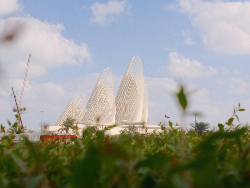 A modern architectural structure resembling sails, surrounded by green foliage under a blue sky with clouds.