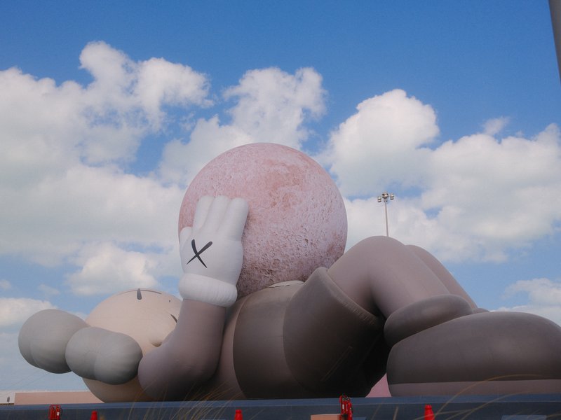 A large cartoonish figure lying on its back with a pink ball, under a blue sky with fluffy clouds.