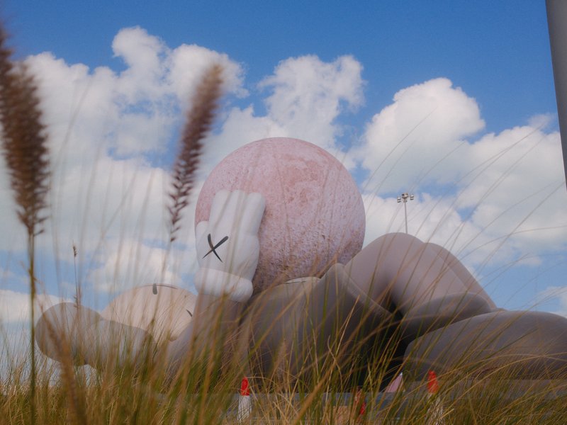 A large pink sculpture resembling a hand holding a ball, surrounded by tall grass and blue sky with clouds.