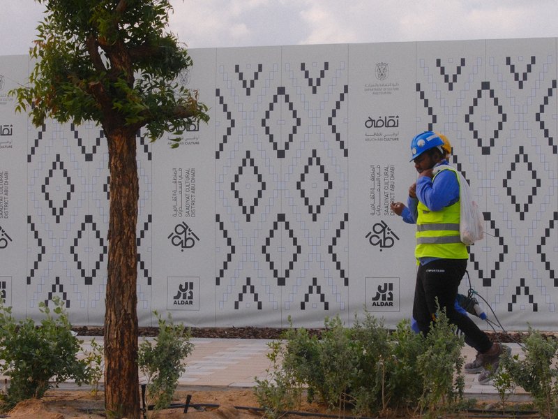 A construction worker in a blue shirt and yellow vest walks past a patterned wall with a tree nearby.