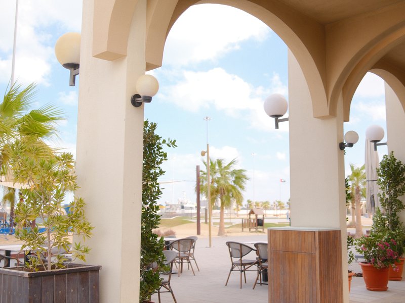 A sunny outdoor cafe scene with arches, palm trees, and blue sky.