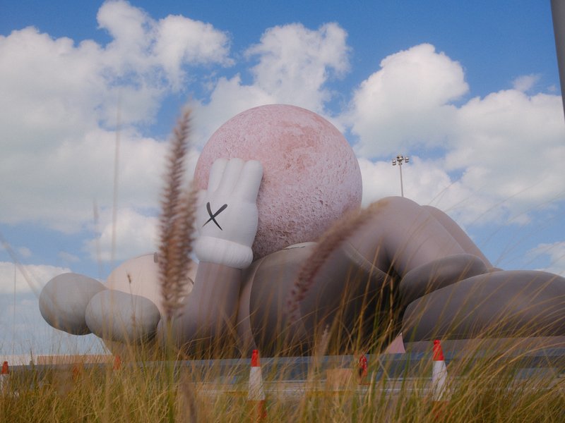 A large, whimsical sculpture of a character lying down with a pink ball head, surrounded by tall grass and blue sky.