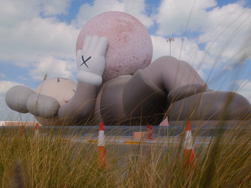 A large inflatable sculpture of a cartoon character lying on its back in a grassy field under a cloudy sky.