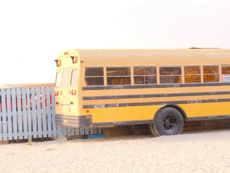A yellow school bus parked beside a wooden fence on a sandy area, with a soft light atmosphere.
