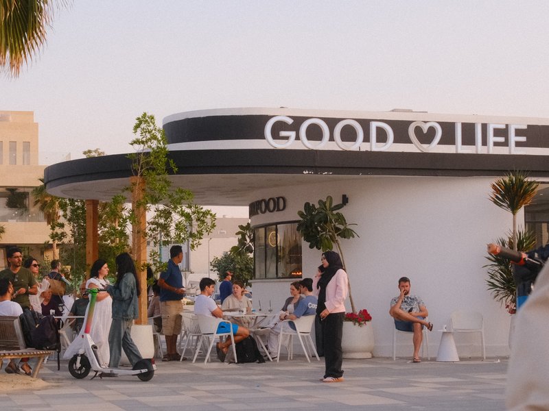 A lively outdoor café scene with people enjoying their time near a sign that reads 'GOOD LIFE'.
