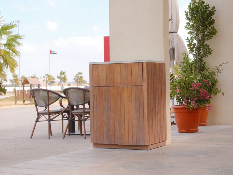 A wooden trash bin beside a table and chairs on a patio with palm trees in the background.