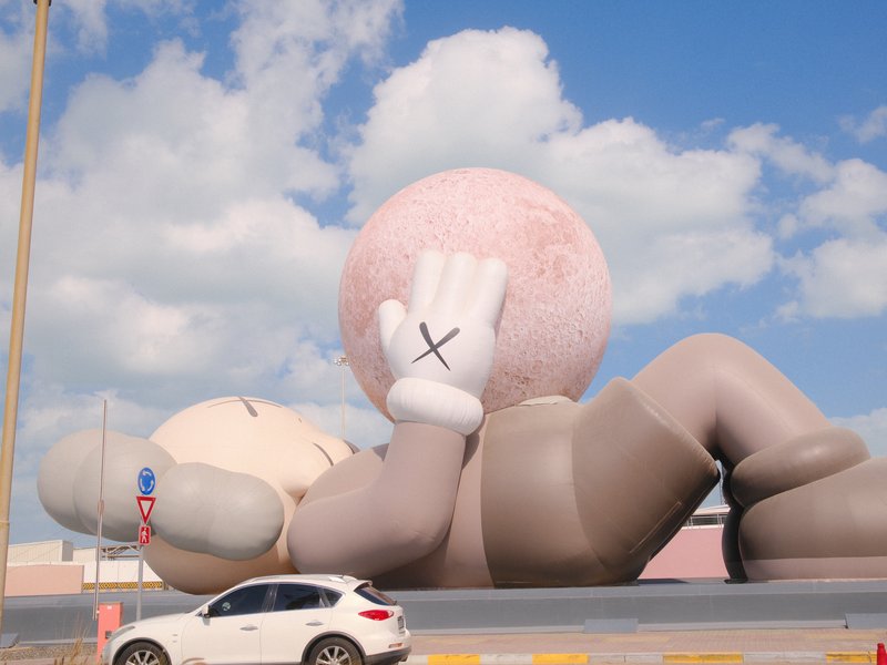 A large cartoonish sculpture of a character lying down with a pink ball, beside a white car.