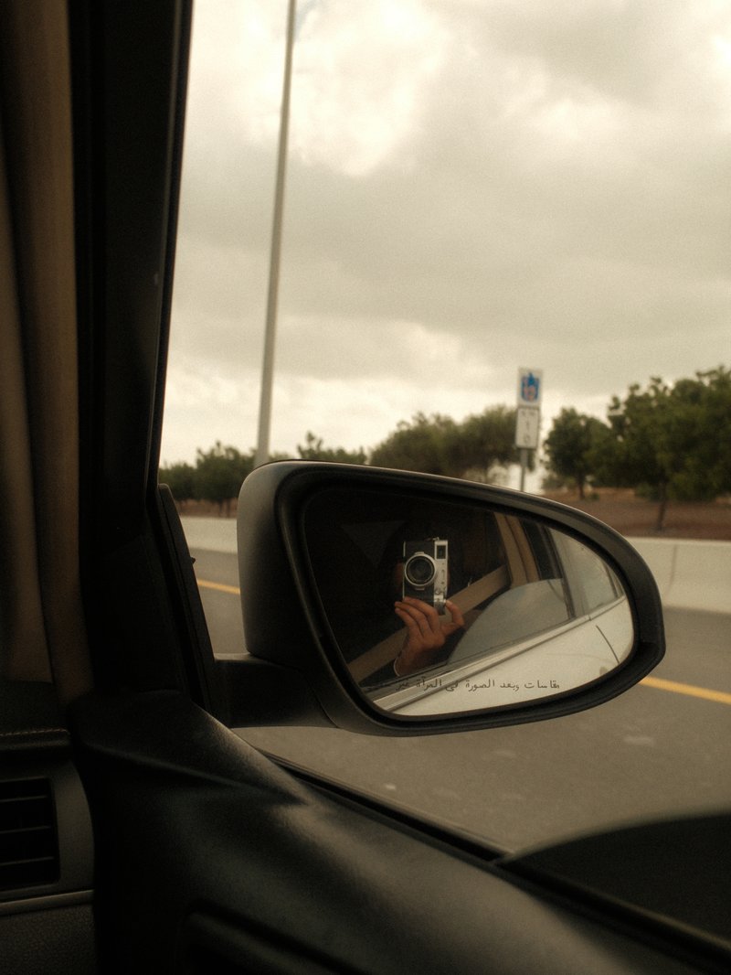 A car's side mirror reflecting a cloudy sky and trees along a highway.