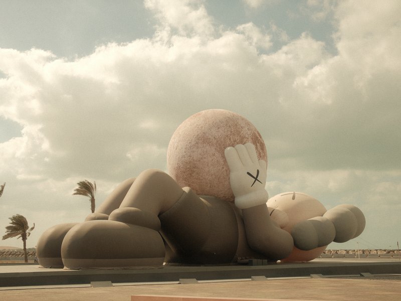 A large inflatable sculpture of a cartoonish figure lying down with a ball, under a cloudy sky.