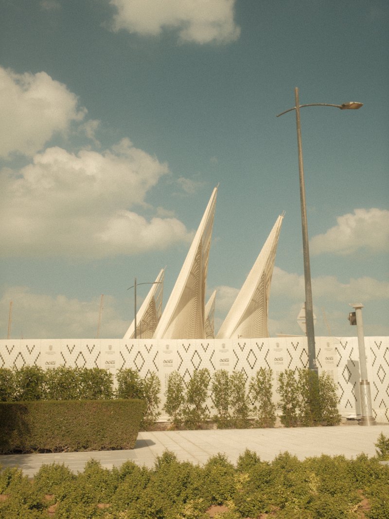 Architectural structure with sharp peaks against a cloudy sky, surrounded by greenery and a white wall.