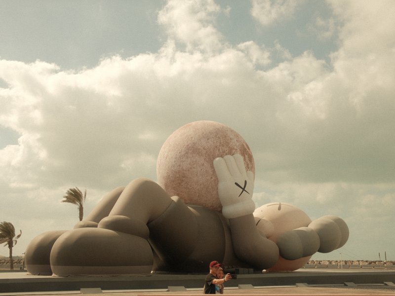 A large cartoonish sculpture of a character lying on the ground, with a person sitting nearby.