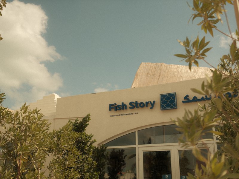 Exterior view of a restaurant named Fish Story with greenery in the foreground and a blue sky.