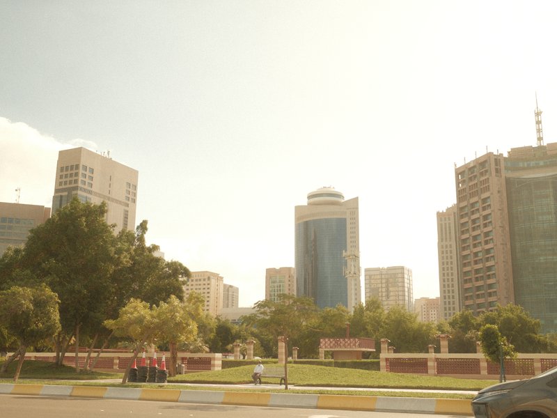 City skyline with modern buildings under a bright sky, featuring trees and a park area in the foreground.