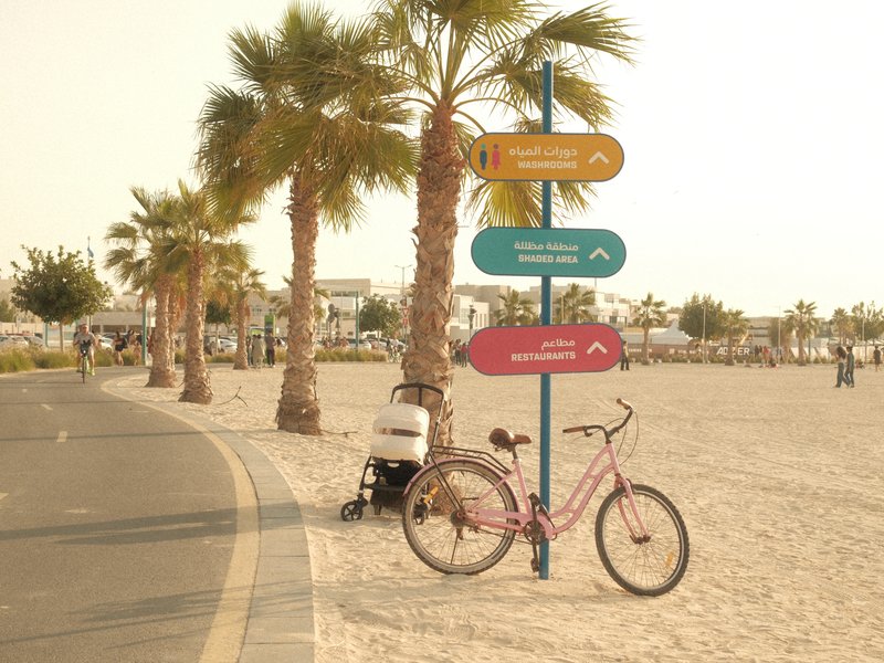 A pink bicycle parked near a signpost on a sandy beach with palm trees in the background.