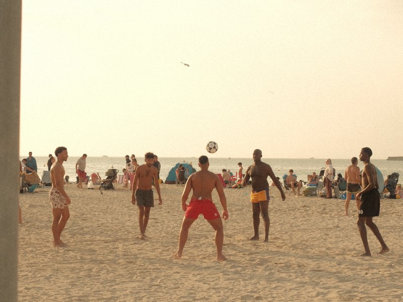 Group of five men playing beach soccer on a sandy beach with people relaxing in the background.