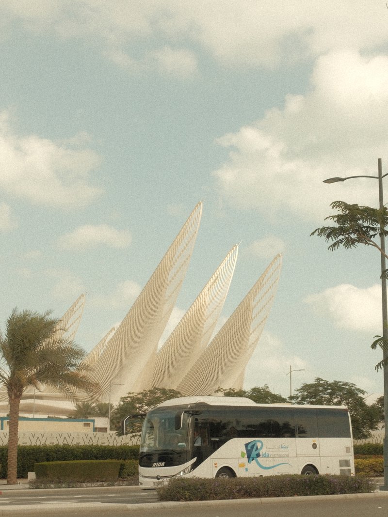 A white bus parked in front of a modern architectural structure with large, pointed sails against a blue sky.