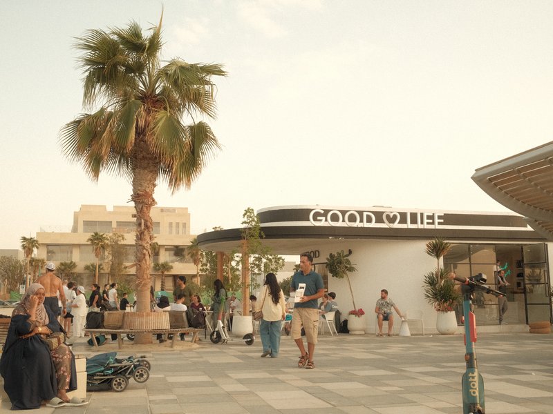 A bustling outdoor plaza with palm trees, people walking, and a modern building in the background.