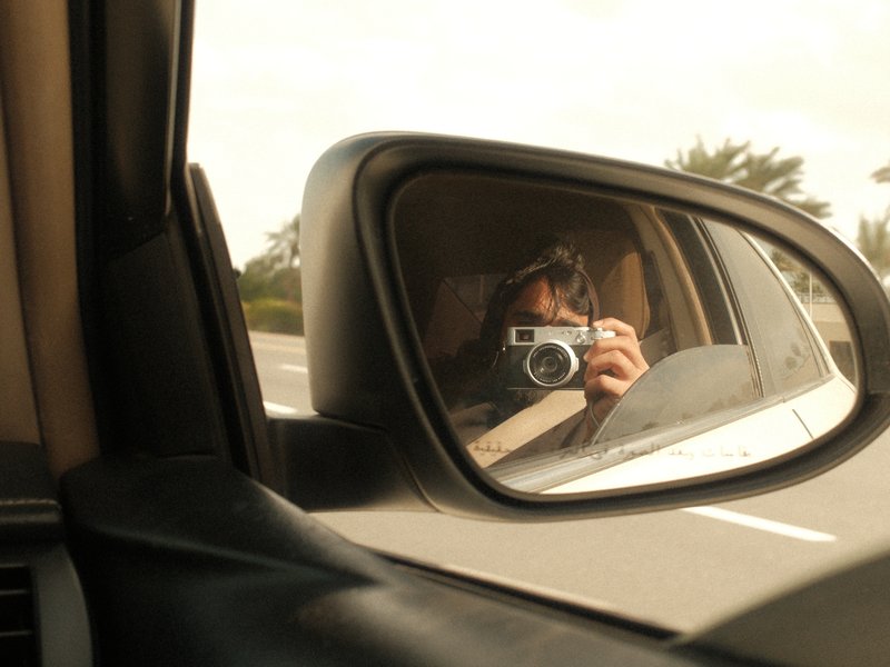 A reflection of a person holding a camera in a car side mirror with a blurred road in the background.