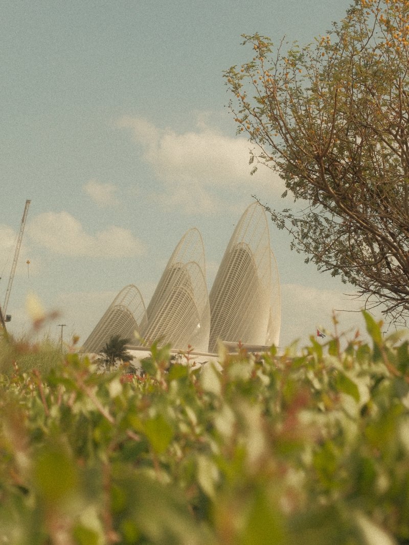 Three modern architectural structures rise above a field of green foliage under a blue sky with clouds.