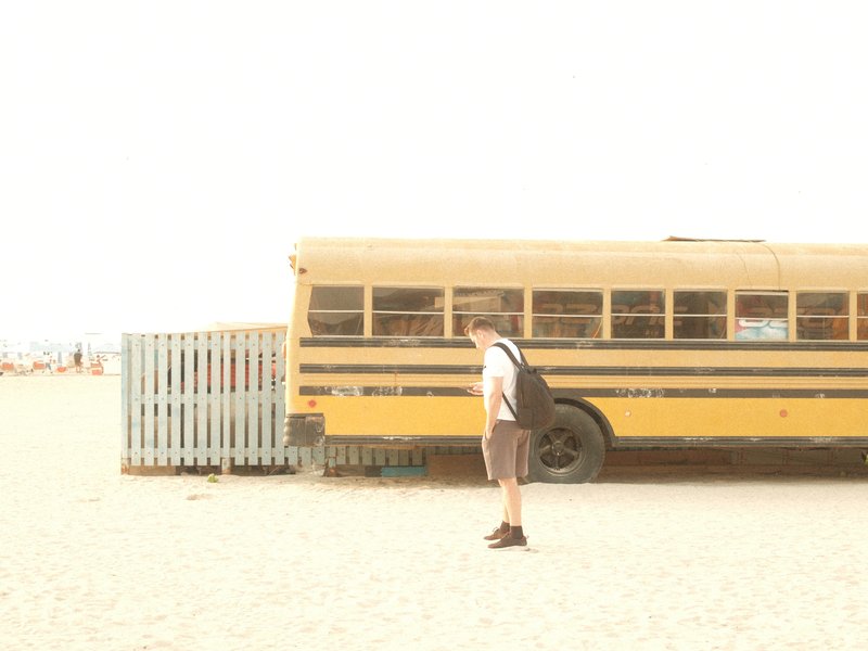 A person with a backpack stands near a yellow school bus on a sandy beach.