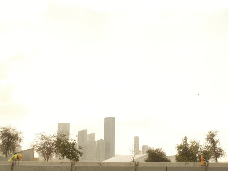 A hazy city skyline with tall buildings and trees in the foreground under a bright sky.