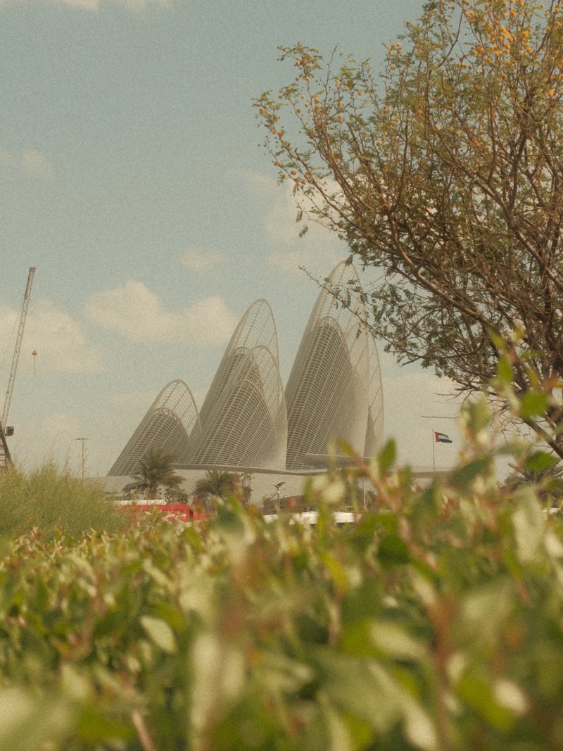 Three modern architectural structures rise behind a green bush under a blue sky with fluffy clouds.