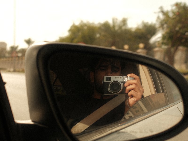 A person holding a camera is reflected in a car's side mirror, with a blurred outdoor background.
