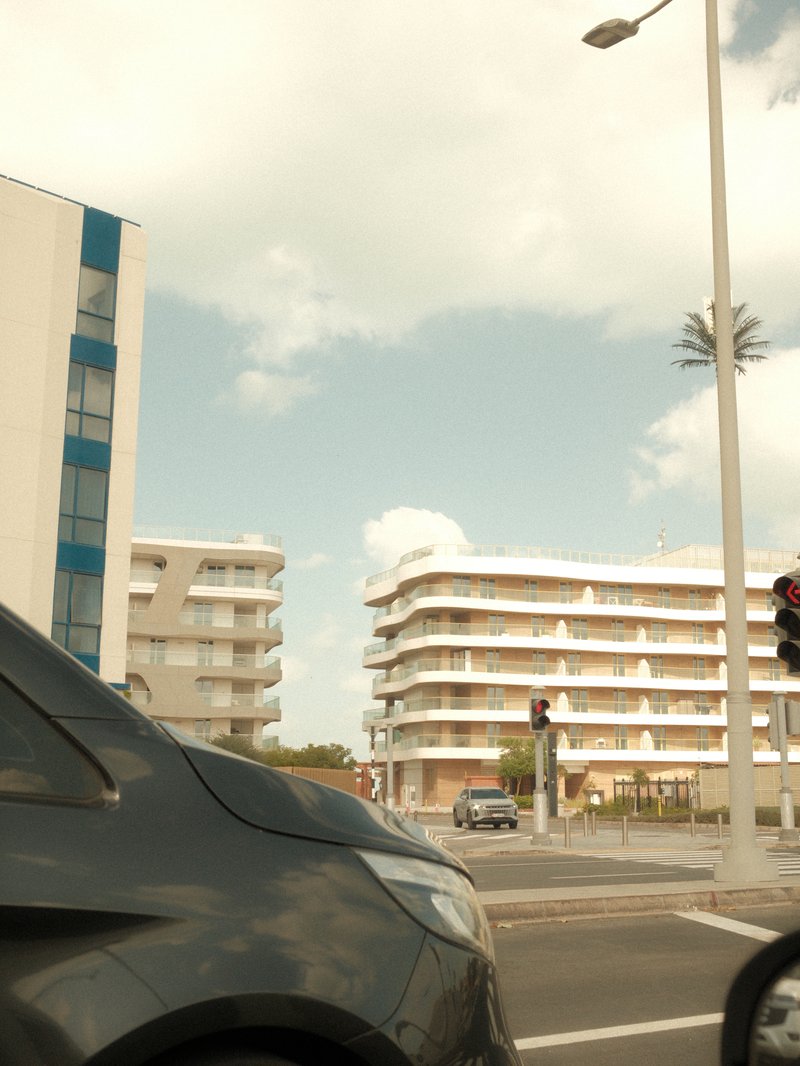 View of modern buildings from a car, with a cloudy sky and palm trees in the background.