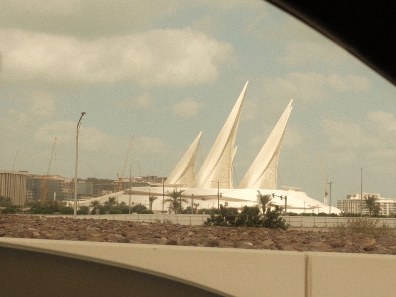 A modern architectural structure with large white sails against a cloudy sky, viewed from a vehicle.