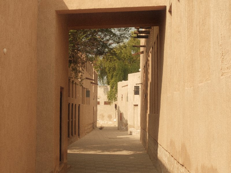 Narrow alleyway between beige buildings with trees in the background under soft light.