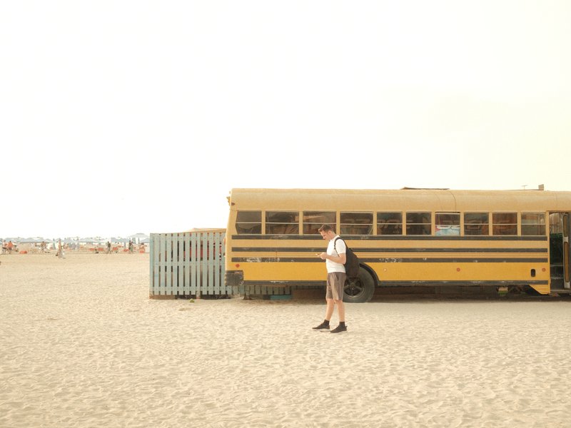 A person stands next to a yellow school bus on a sandy beach with a hazy background.