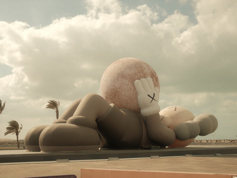 A large cartoonish sculpture of a character lying on its back, holding a sphere, with palm trees in the background.