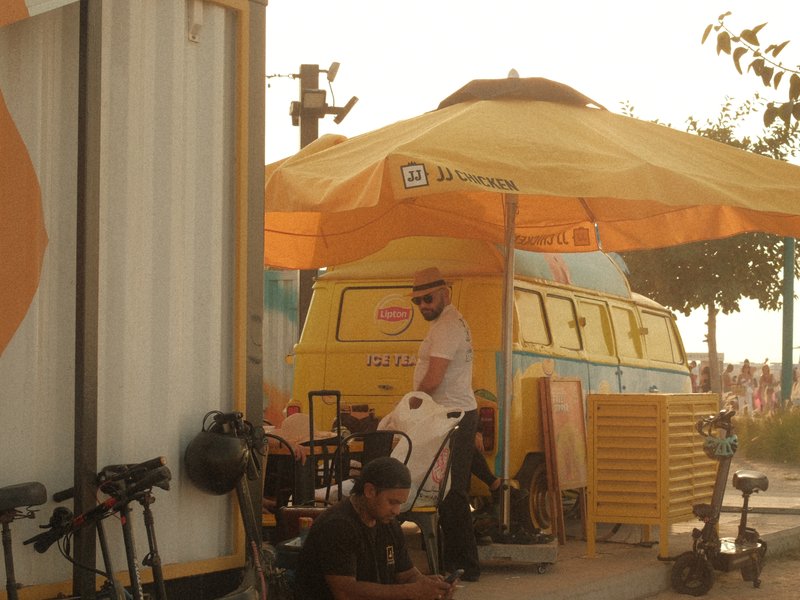 A man in a white shirt stands near a colorful food truck under a large yellow umbrella.