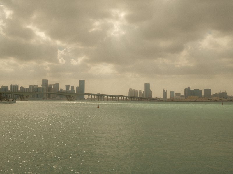A city skyline with a bridge over water under a cloudy sky, light reflecting on the water surface.