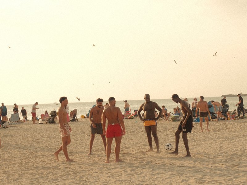 Group of men playing soccer on a beach with people lounging in the background under a hazy sky.