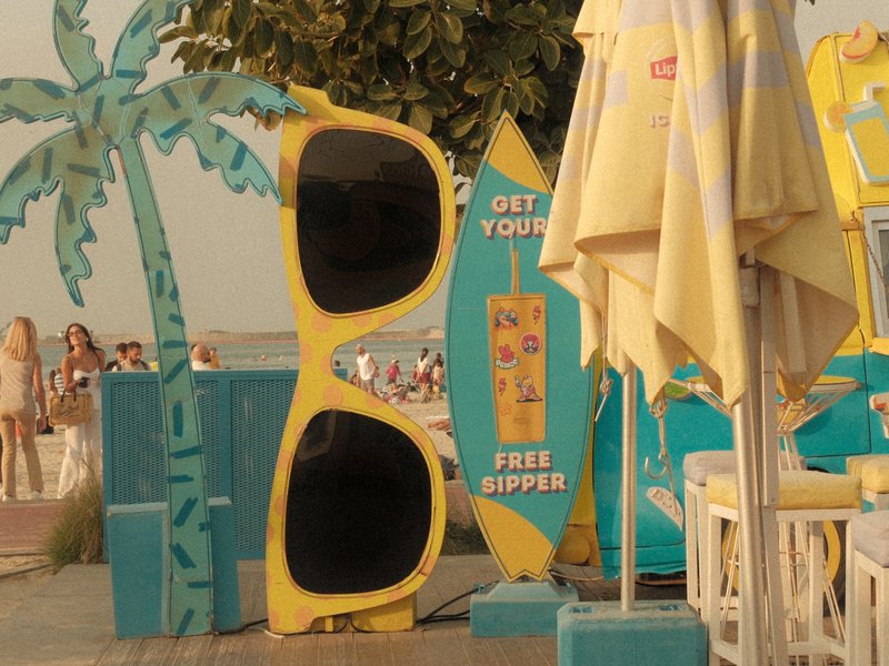 Colorful beach scene featuring oversized sunglasses and surfboards with people in the background.