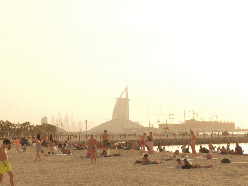 Beach scene with people lounging on sand, a windmill structure in the background under a hazy sky.