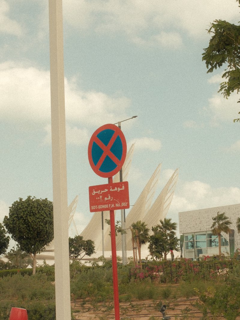 A no parking sign in a landscaped area with modern architecture in the background under a cloudy sky.
